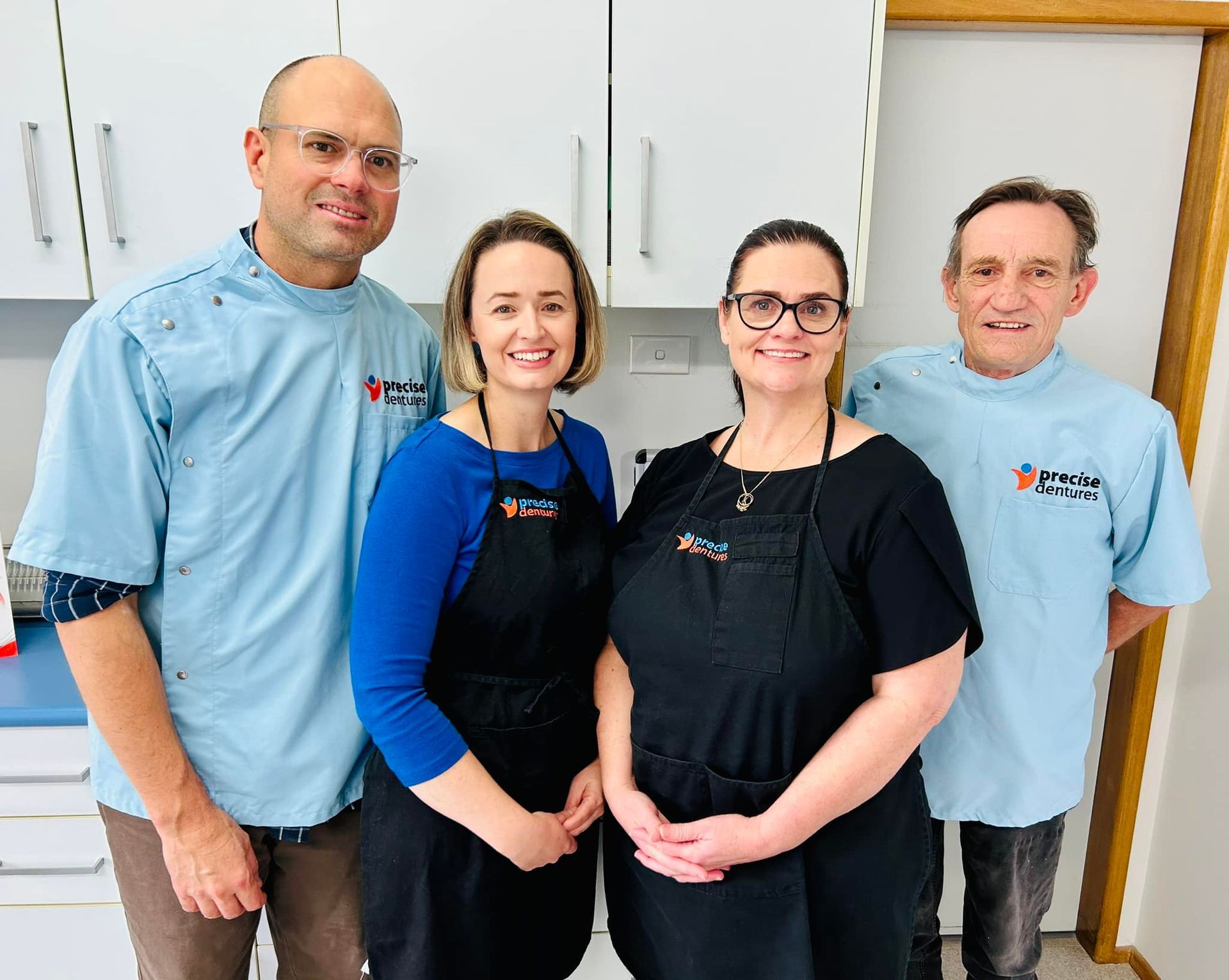 Four Clinic Staff in Blue and Black Uniforms Posing Together in a Bright Hallway — Precise Dentures in Berserker, QLD
