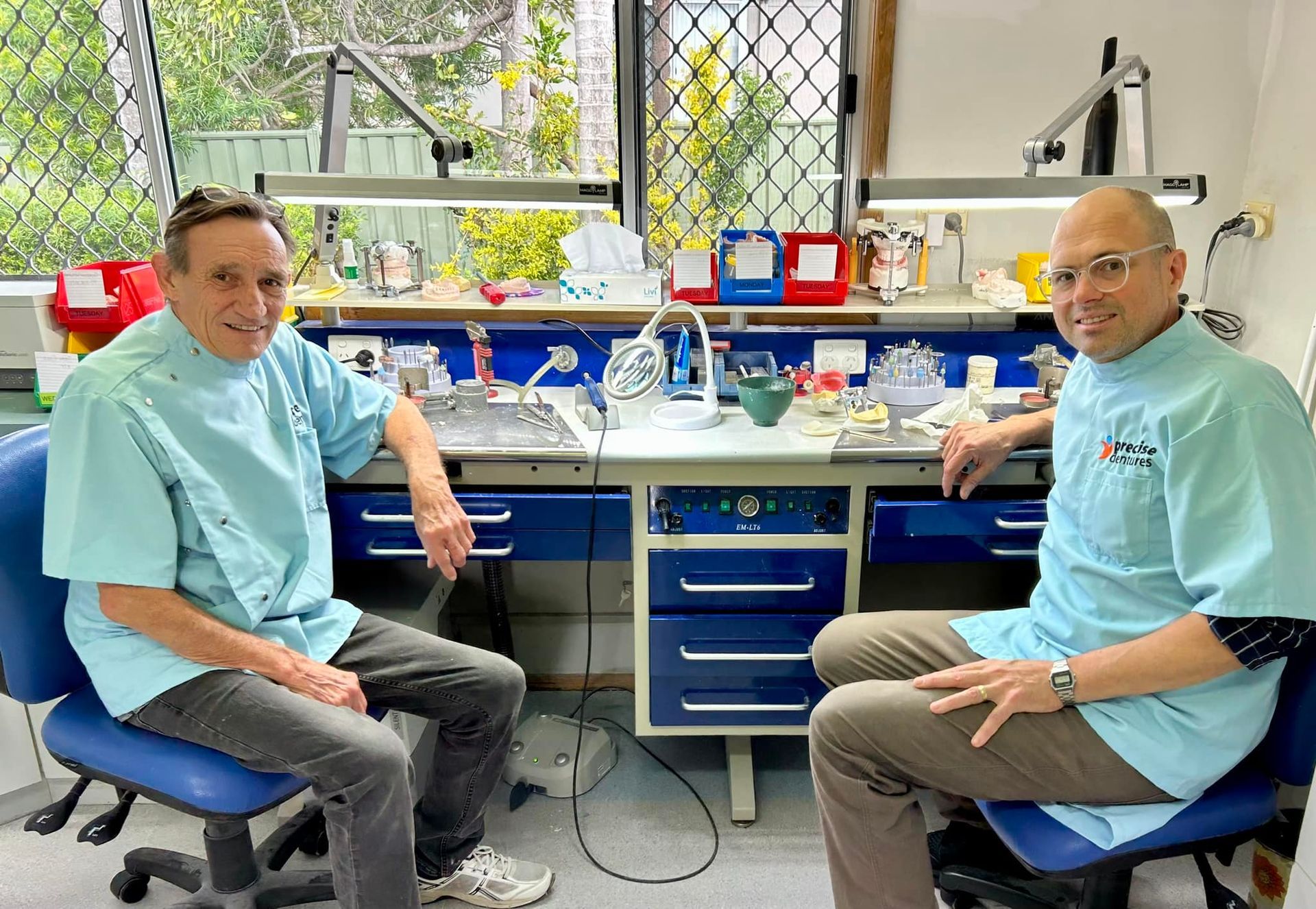 Two Clinic Staff Sitting in a Bright Lab, Smiling Beside a Workbench — Precise Dentures in Berserker, QLD