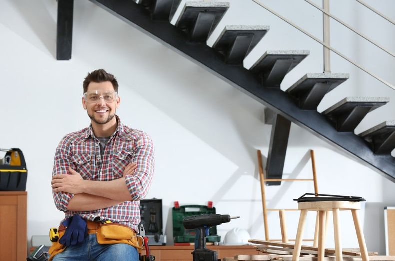 Smiling person with folded arms in a workshop beneath a staircase, with tools and wooden stools nearby