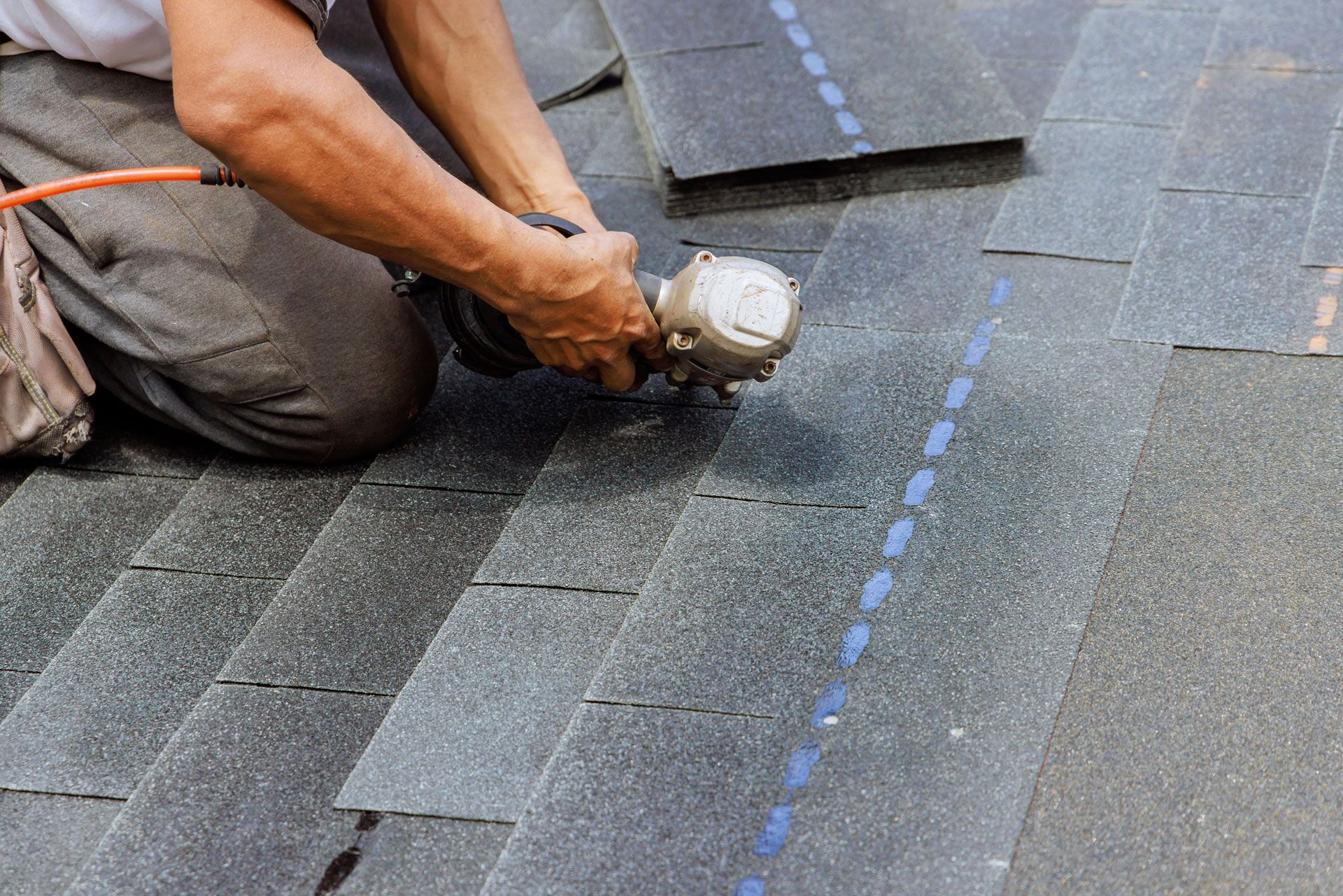 Roofer kneeling, using a nail gun on dark shingles to repair a roof.