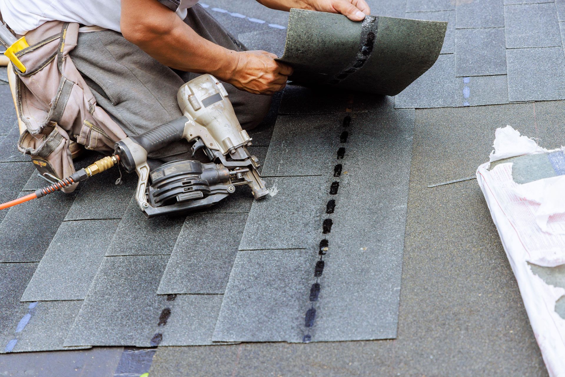 Roofer installing dark gray asphalt shingles with a nail gun on a roof.