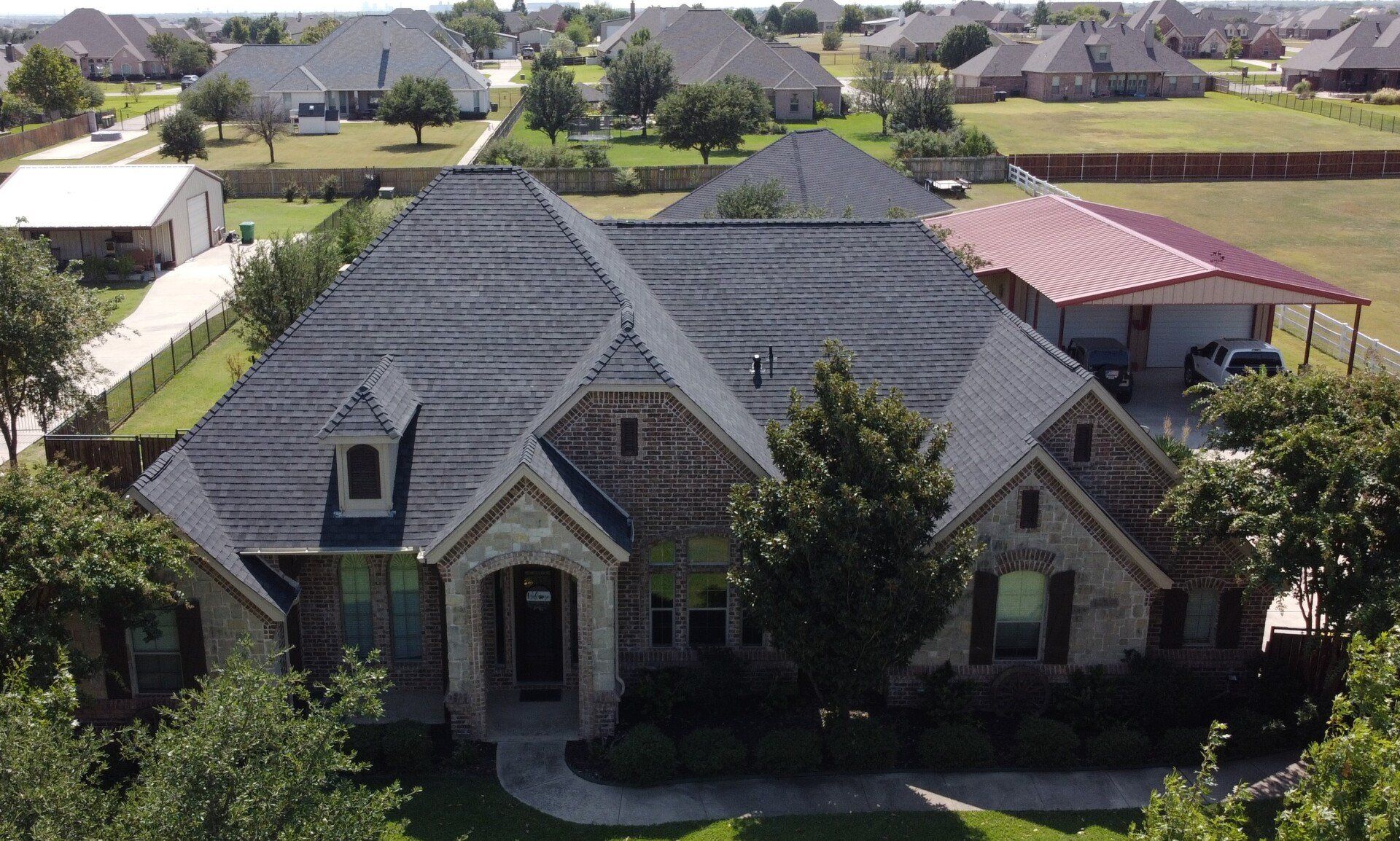Aerial view of a large gray suburban house with a steep roof and front porch, surrounded by trees and neighboring homes