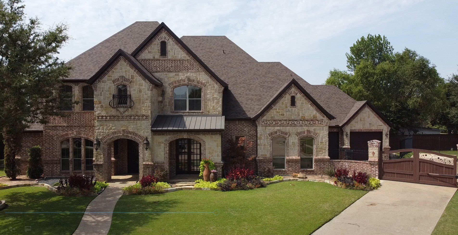 Large stone suburban house with manicured lawn, flower beds, and a driveway under a cloudy sky