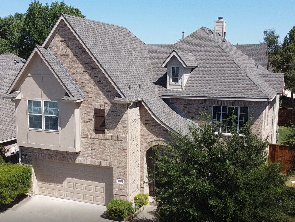 Stone suburban house with gray roof, garage, and front trees under bright daylight