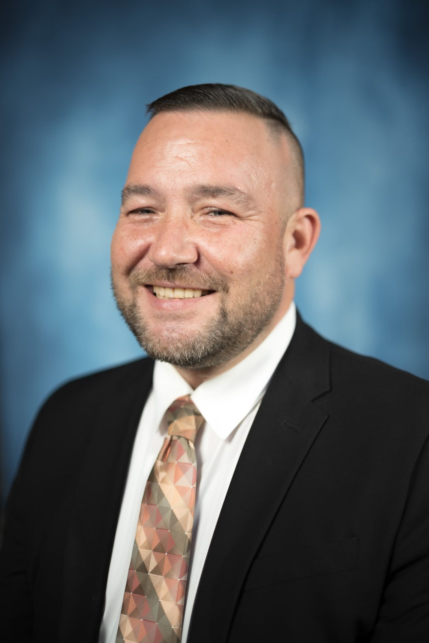 Man in suit smiles. He has a shaved head, stubble, and a patterned tie. Blue background.