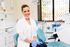 Dentist smiling in a dental office, wearing gloves and a white coat, seated in a dental chair.