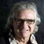 Man with silver hair, glasses, and a plaid shirt, smiling at the camera. Dark background.