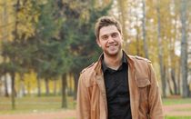 Man in brown jacket smiles at camera in an autumn park.