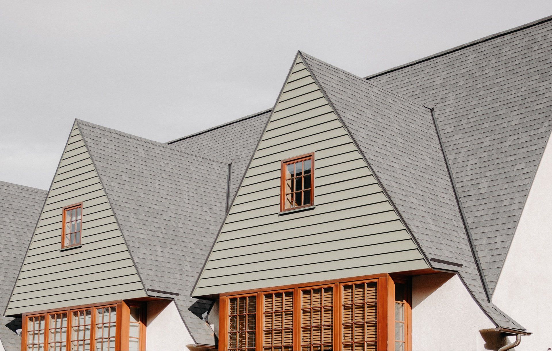 Gray-shingled house roof with steep dormers and wooden windows under a cloudy sky