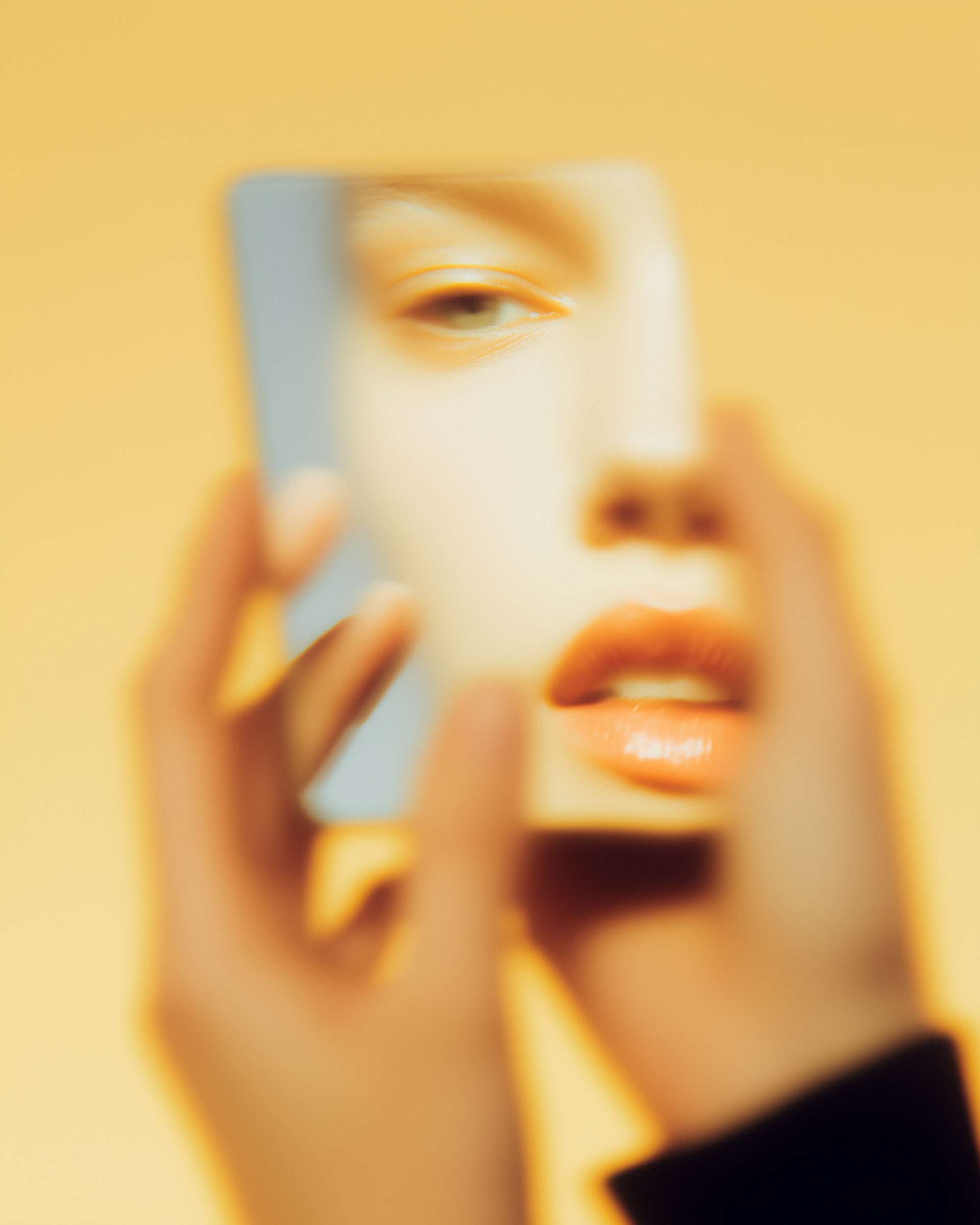 A blurry, close-up photo shows hands holding a small mirror reflecting part of a face against a solid yellow background.