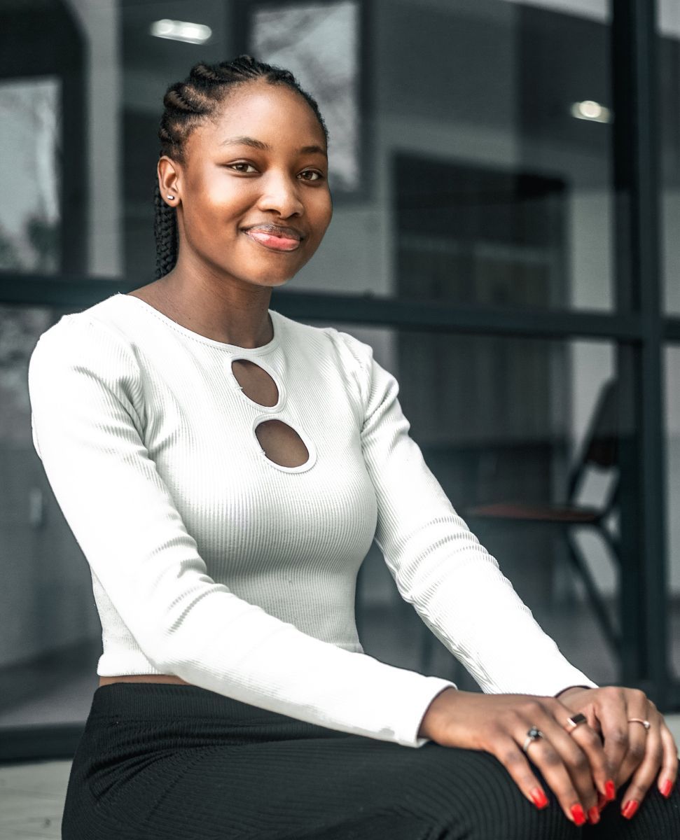 Woman in white top with keyhole details, sitting with hands clasped, smiling. Modern building background.