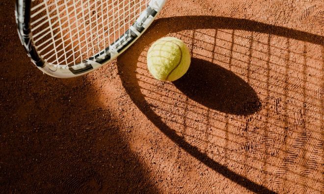 Tennis racket and ball on a clay court, casting a shadow.
