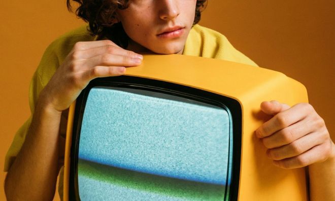 Person leaning on a vintage yellow television, looking thoughtful against a yellow background.