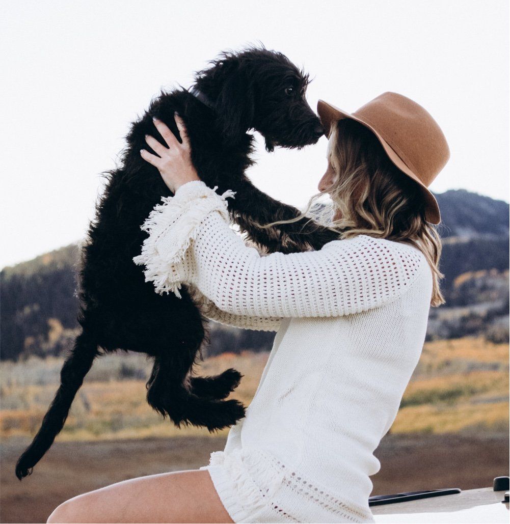 Een vrouw met een beige hoed tilt een zwarte hond op in een grasveld met bergen op de achtergrond.