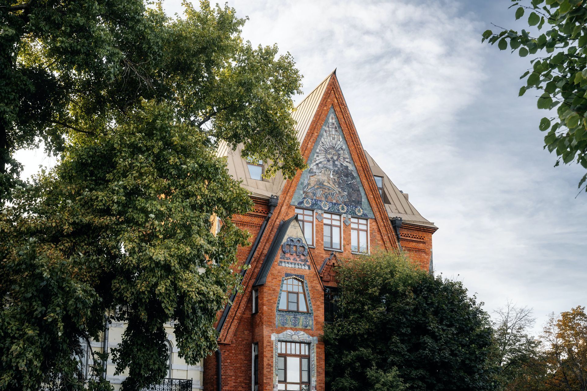 Red-brick Victorian house with steep gabled roof, partly framed by leafy trees under a cloudy sky