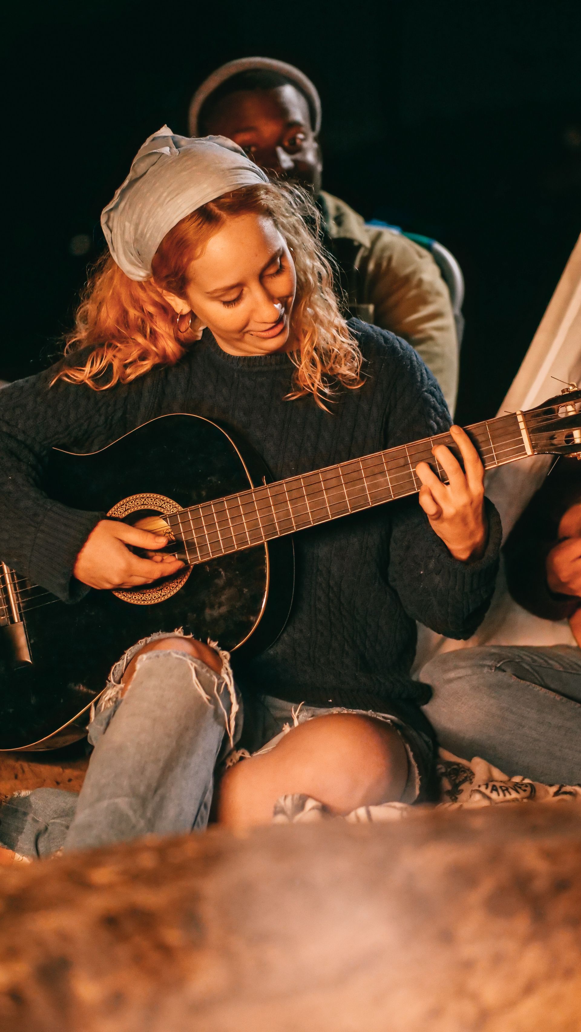 Woman playing acoustic guitar by a campfire, wearing a knit hat and sweater.