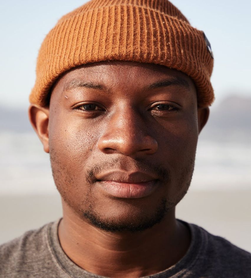 Portrait of a man in a brown knit beanie, facing the camera on a beach with a blurred shoreline behind him