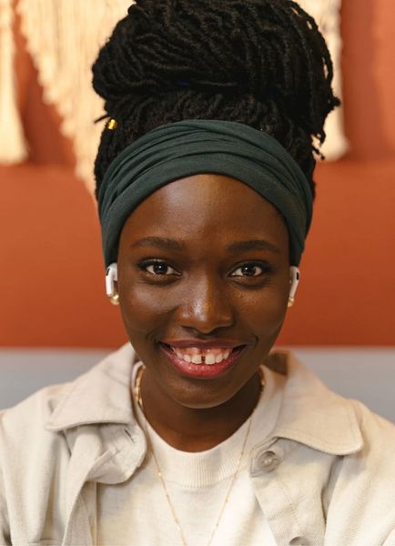 Smiling person with a dark headwrap and white shirt, seated indoors against a warm-toned background