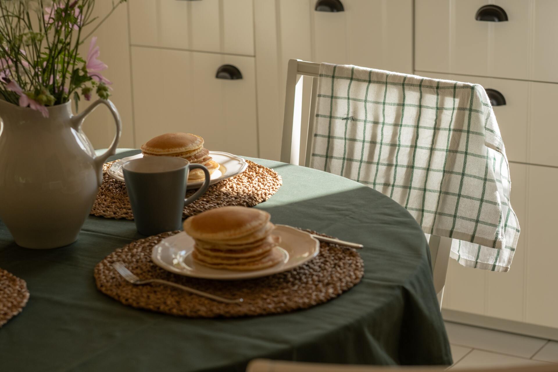 Stack of pancakes on a plate, on a table set for breakfast with a pitcher of flowers.