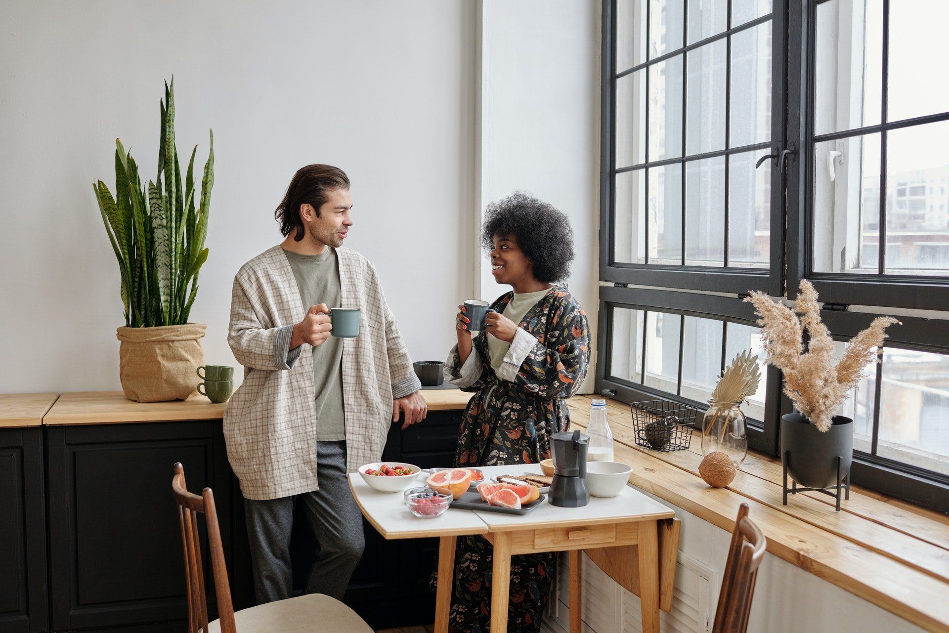 Couple in robes chatting near a window, holding mugs; breakfast on a table.