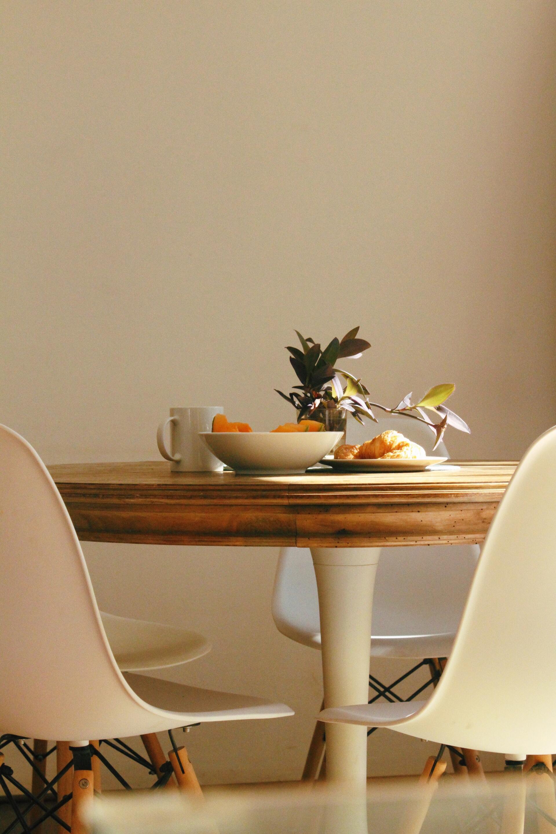 Wooden dining table with white chairs, bowl of food, mug, and plant. Bright sunlight.