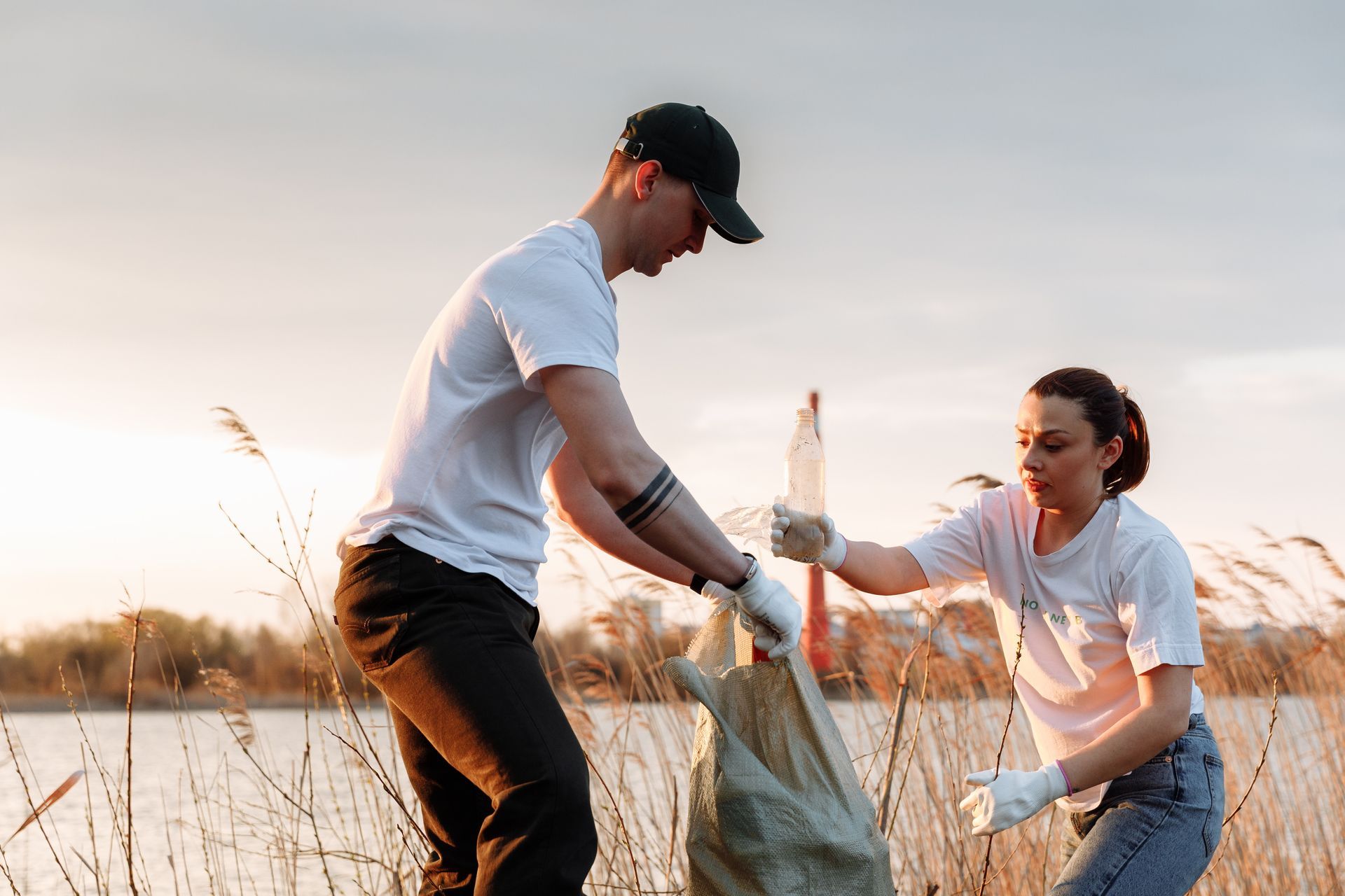 Two people collecting trash near a body of water, wearing white shirts and gloves.