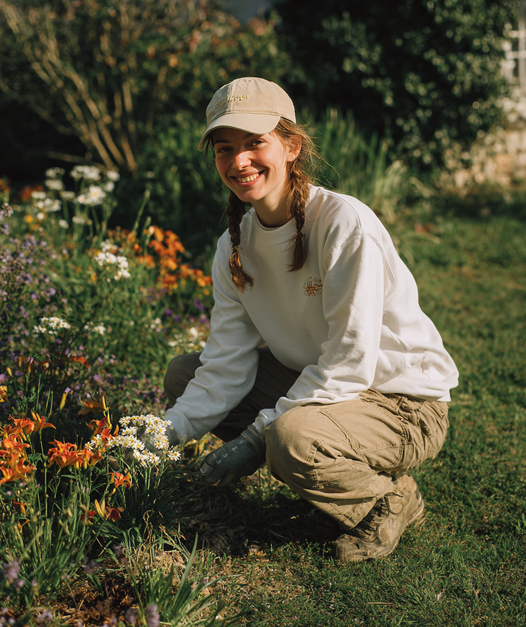 Person kneeling in a sunlit flower garden, smiling and holding white blossoms, wearing a cap and cream sweater