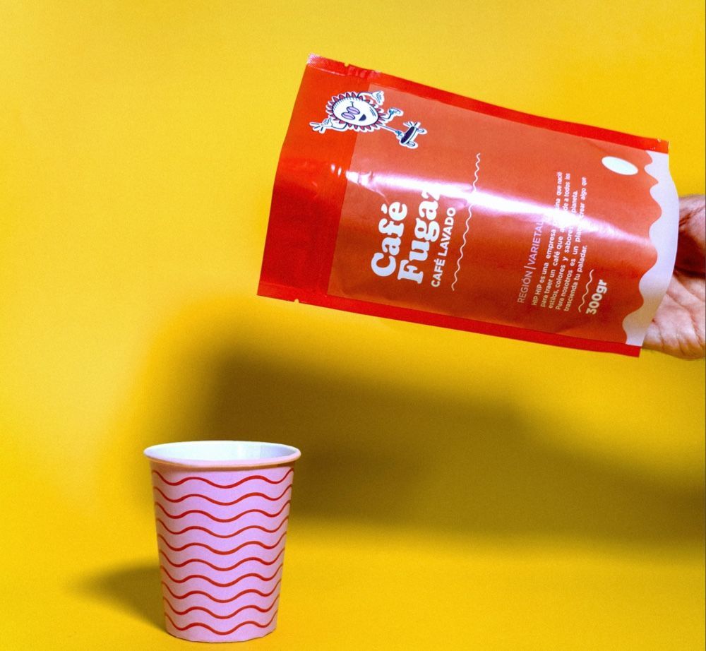 Person pouring coffee from a red package into a pink paper cup against a yellow background.