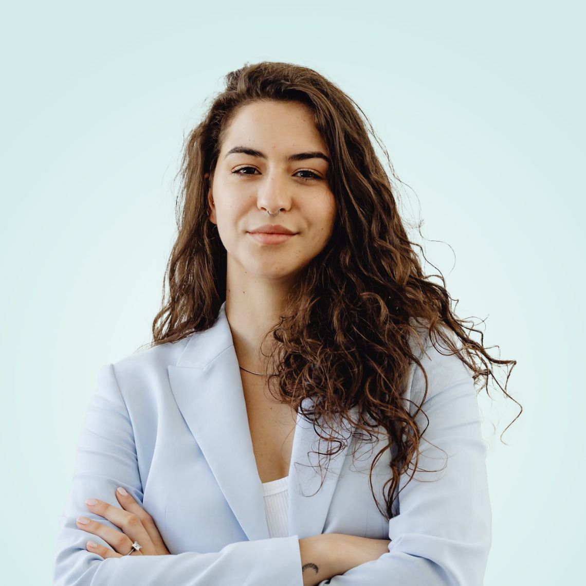 Mujer con cabello rizado, vestida con un blazer azul claro, de pie con los brazos cruzados sobre un fondo azul pálido.