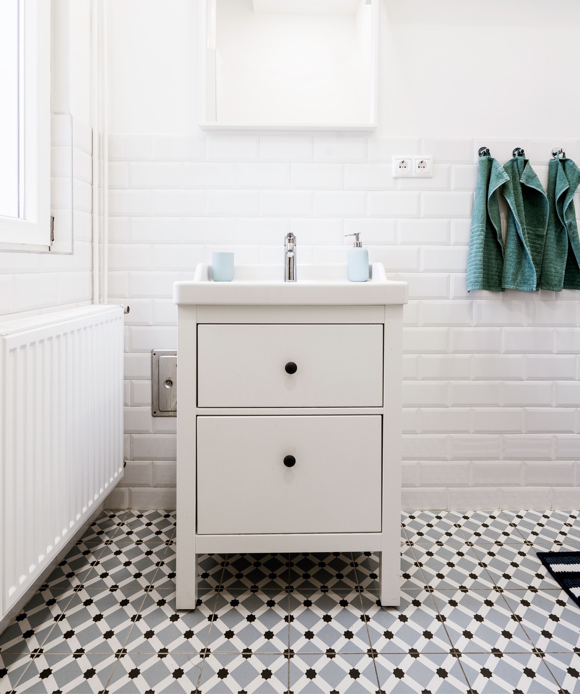 Bright white bathroom vanity with sink and mirror, black-and-white patterned tile floor, and towels on wall hooks