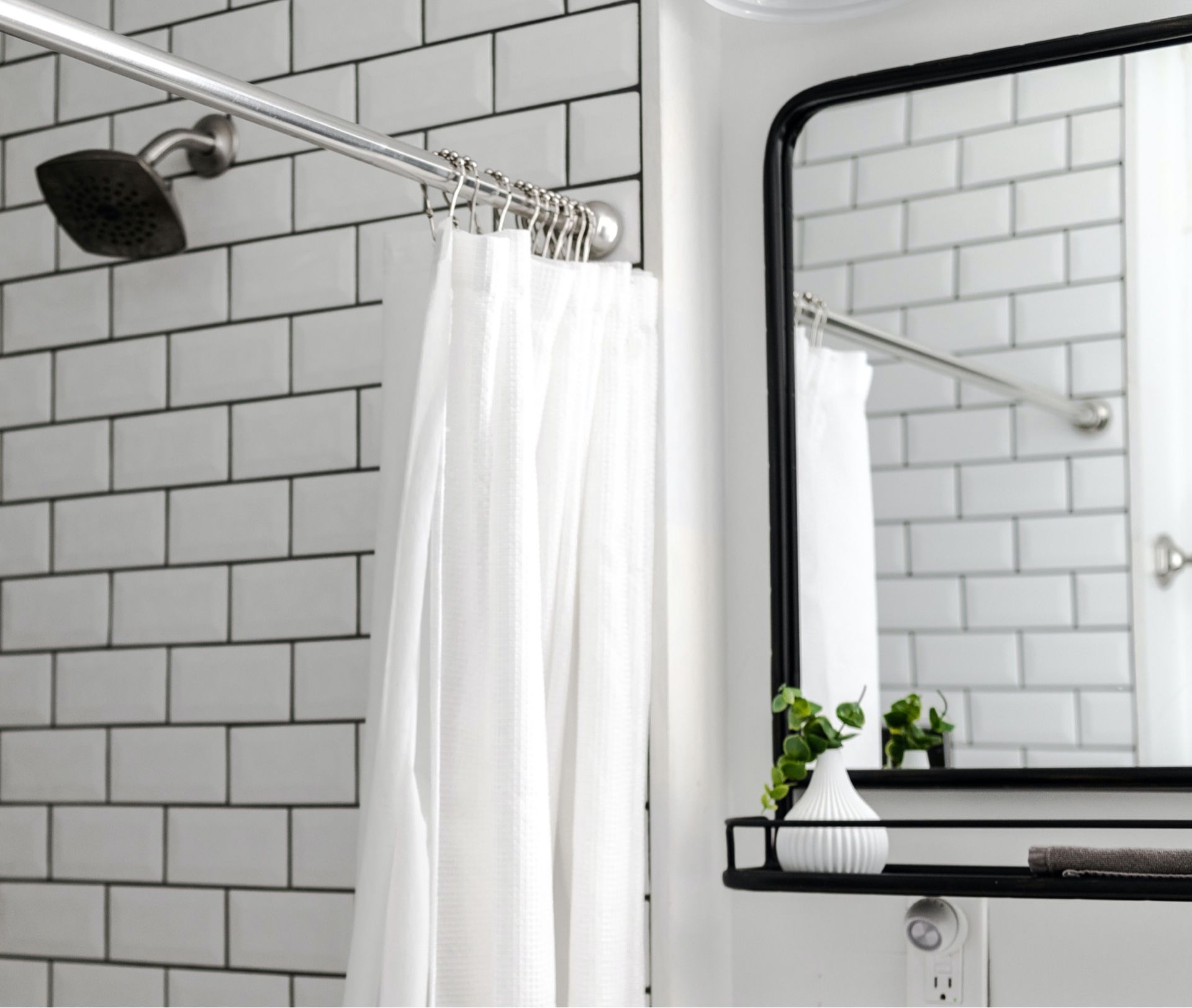 Black showerhead and white curtain in a white subway-tile bathroom, with a mirror and small shelf.