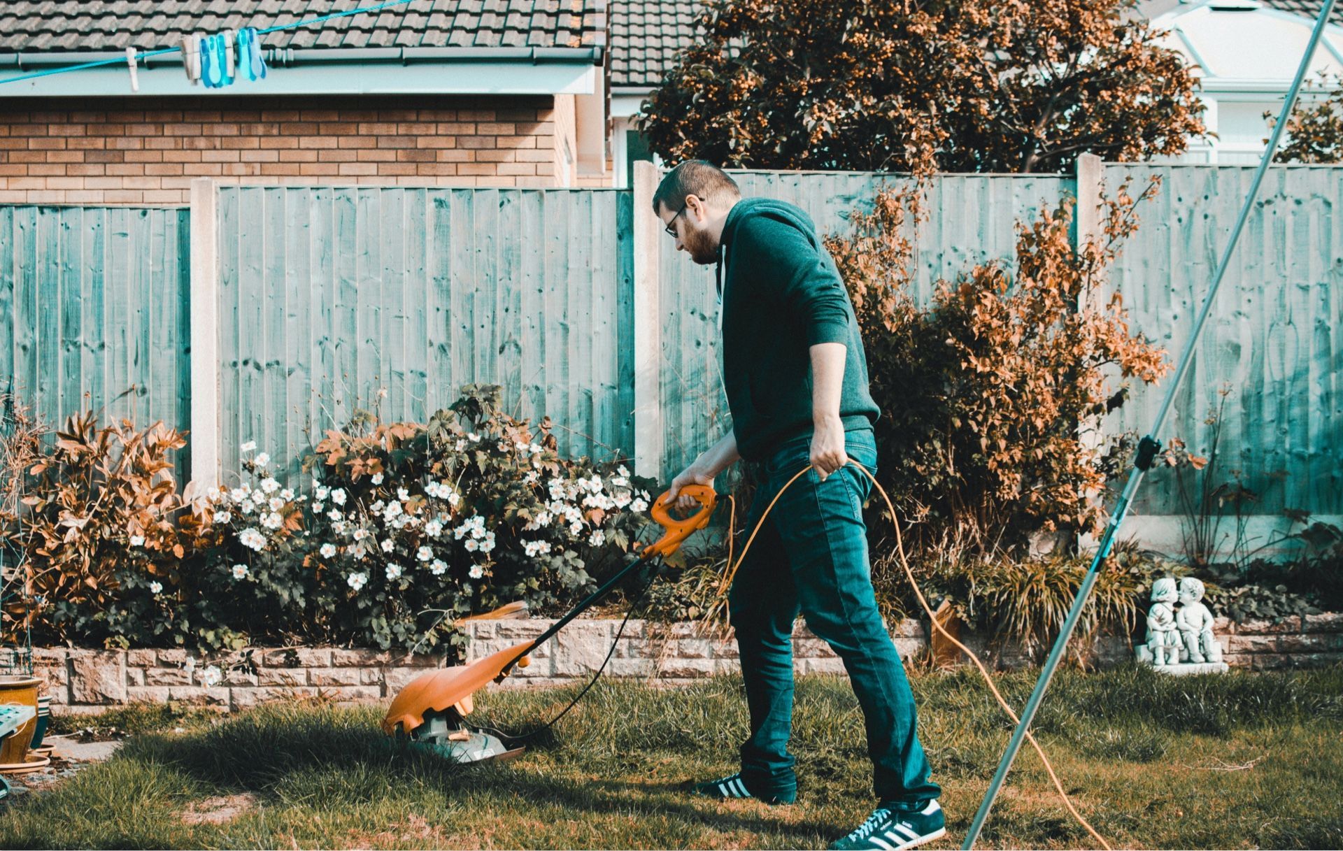 Person mowing a grassy backyard beside a wooden fence and flowers.
