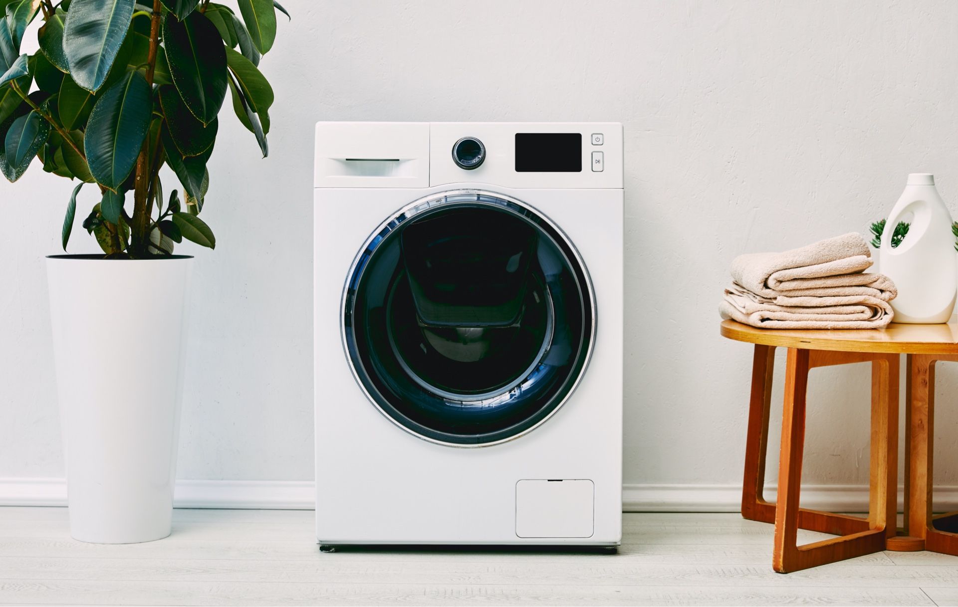 White front-load washing machine in a bright room beside a potted plant and folded towels on a stool.