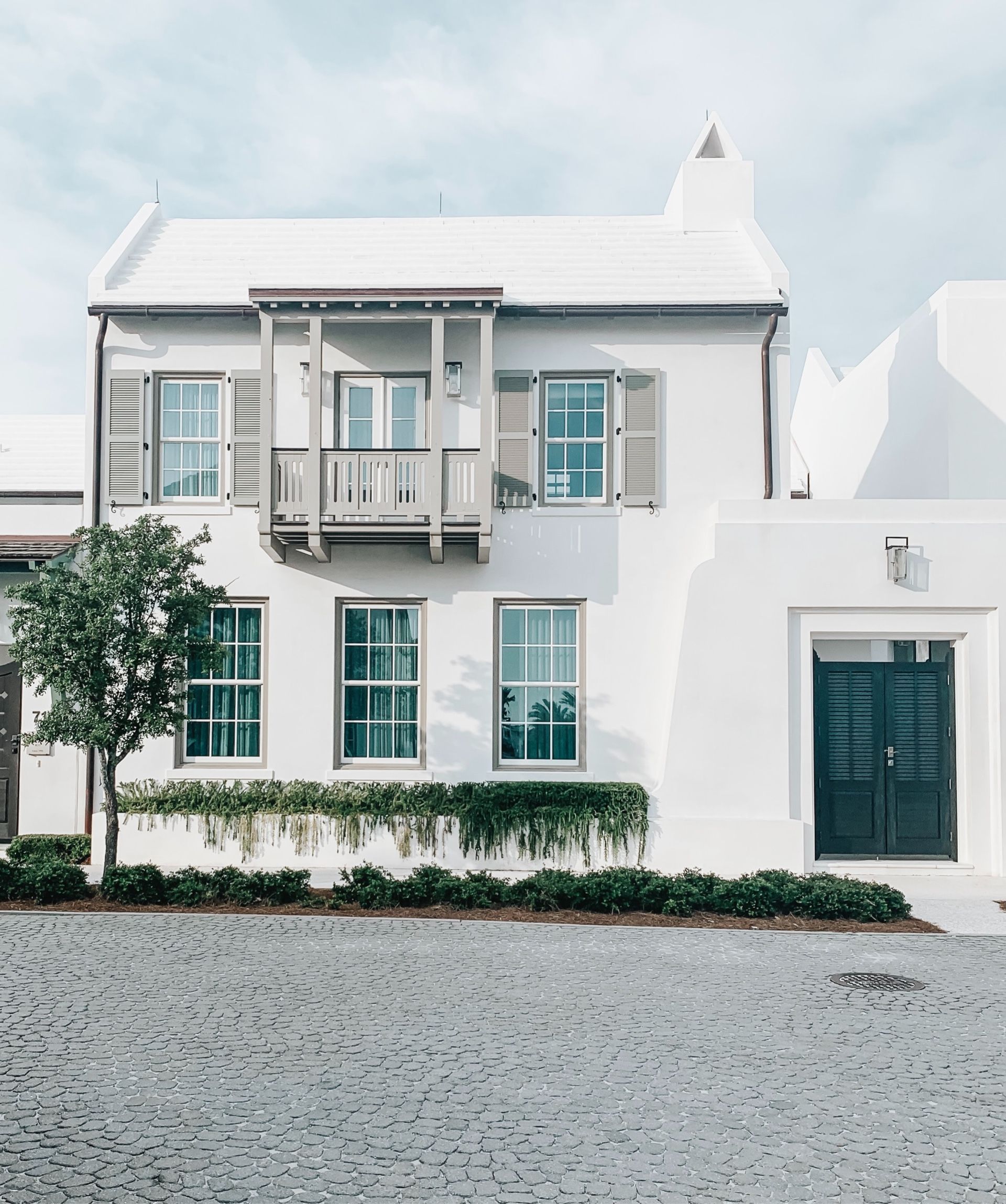 White two-story house with teal windows, balcony, dark green door, and gravel driveway