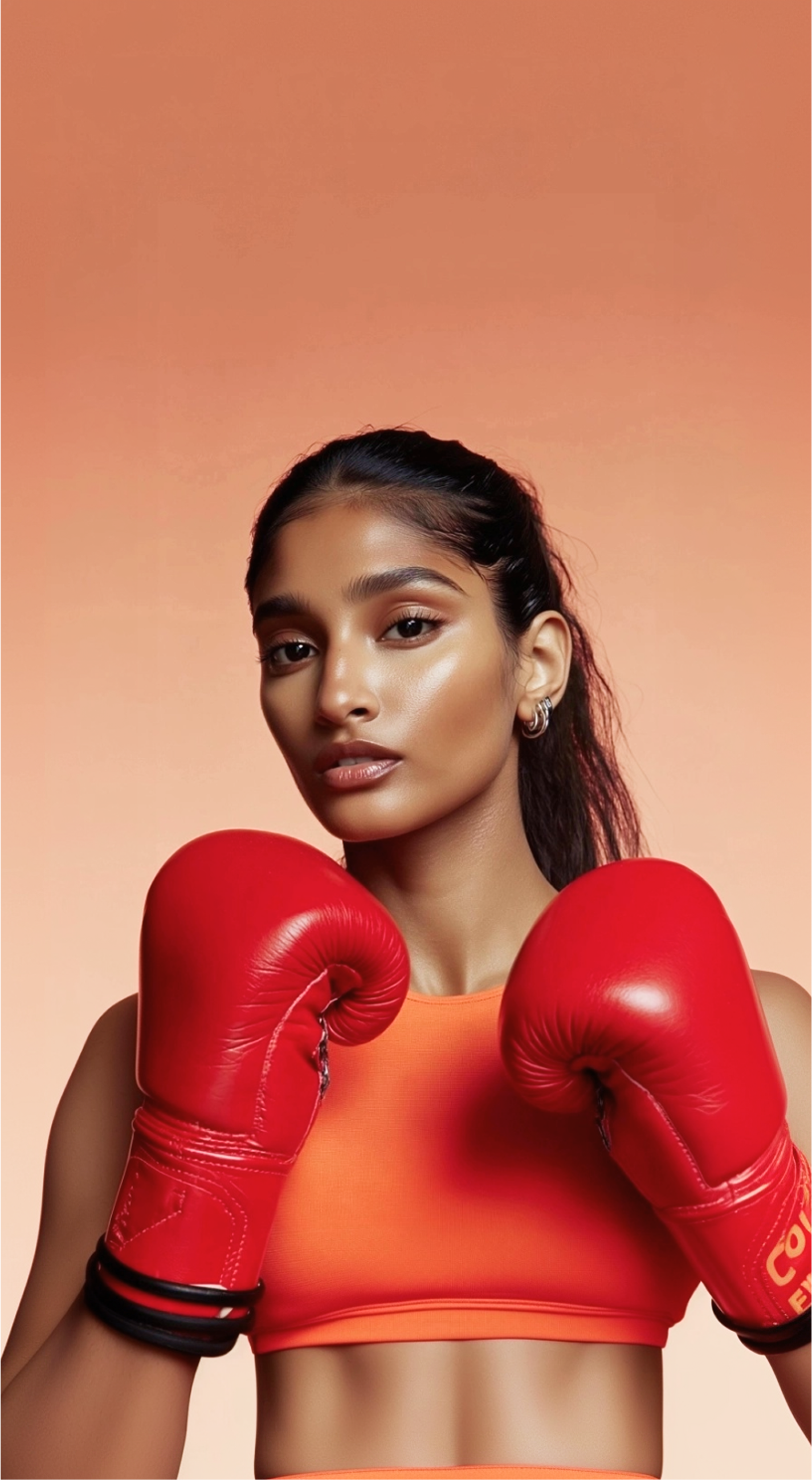 Een vrouw in een oranje sportbeha en rode bokshandschoenen poseert voor een oranje achtergrond.