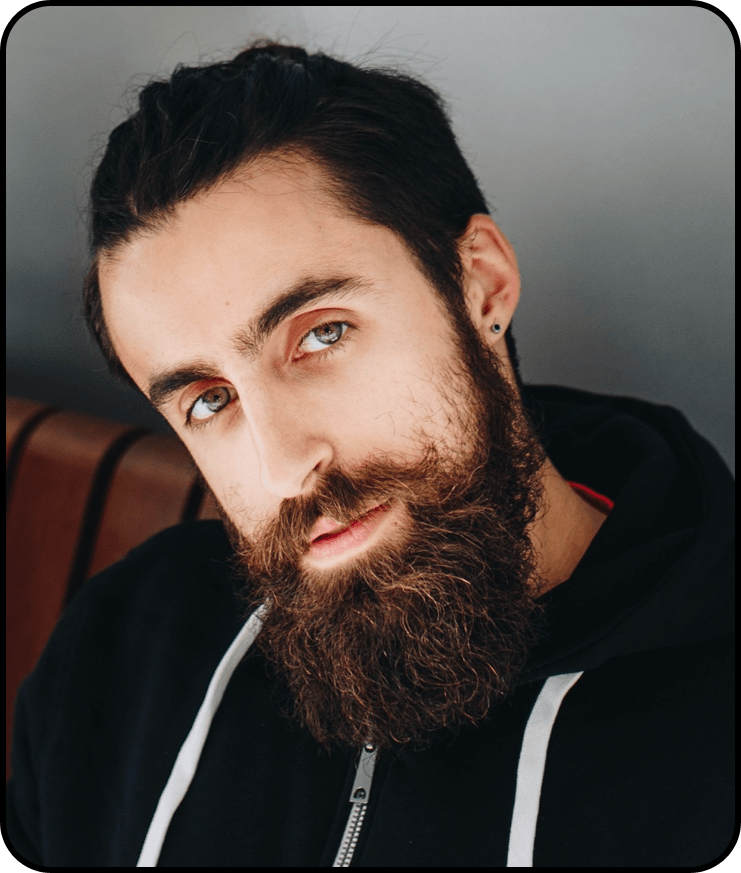 Een persoon met donker haar en een volle baard kijkt voor zich uit, gekleed in een zwarte hoodie met witte trekkoorden.