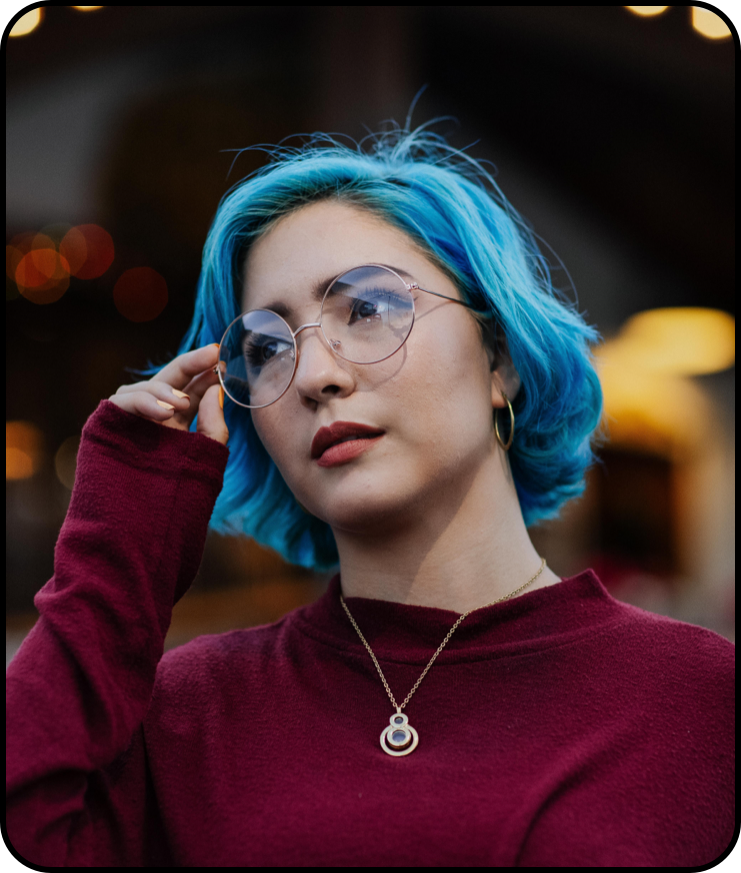 Een persoon met kort, felblauw haar en een ronde bril, gekleed in een donkerrode trui en met een gouden hanger aan een ketting.