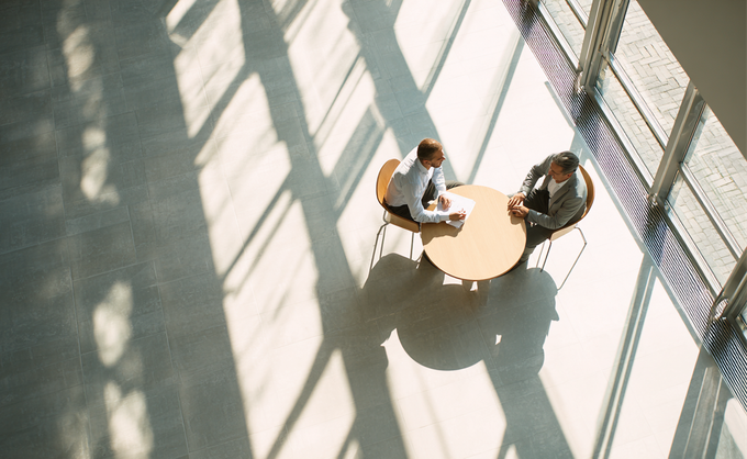 Two people sit at a small round table in a sunlit lobby with long window shadows.