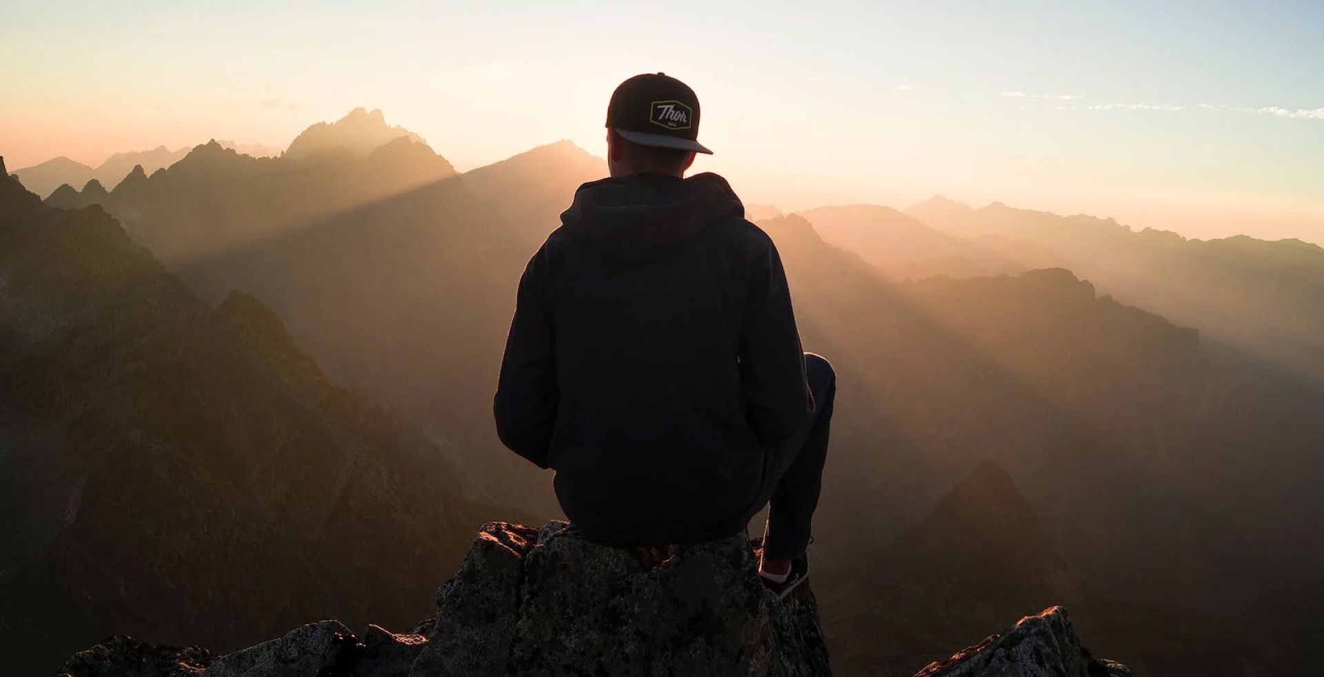Person in hoodie and cap sits on a mountain peak, looking at a sunrise over a range of mountains.