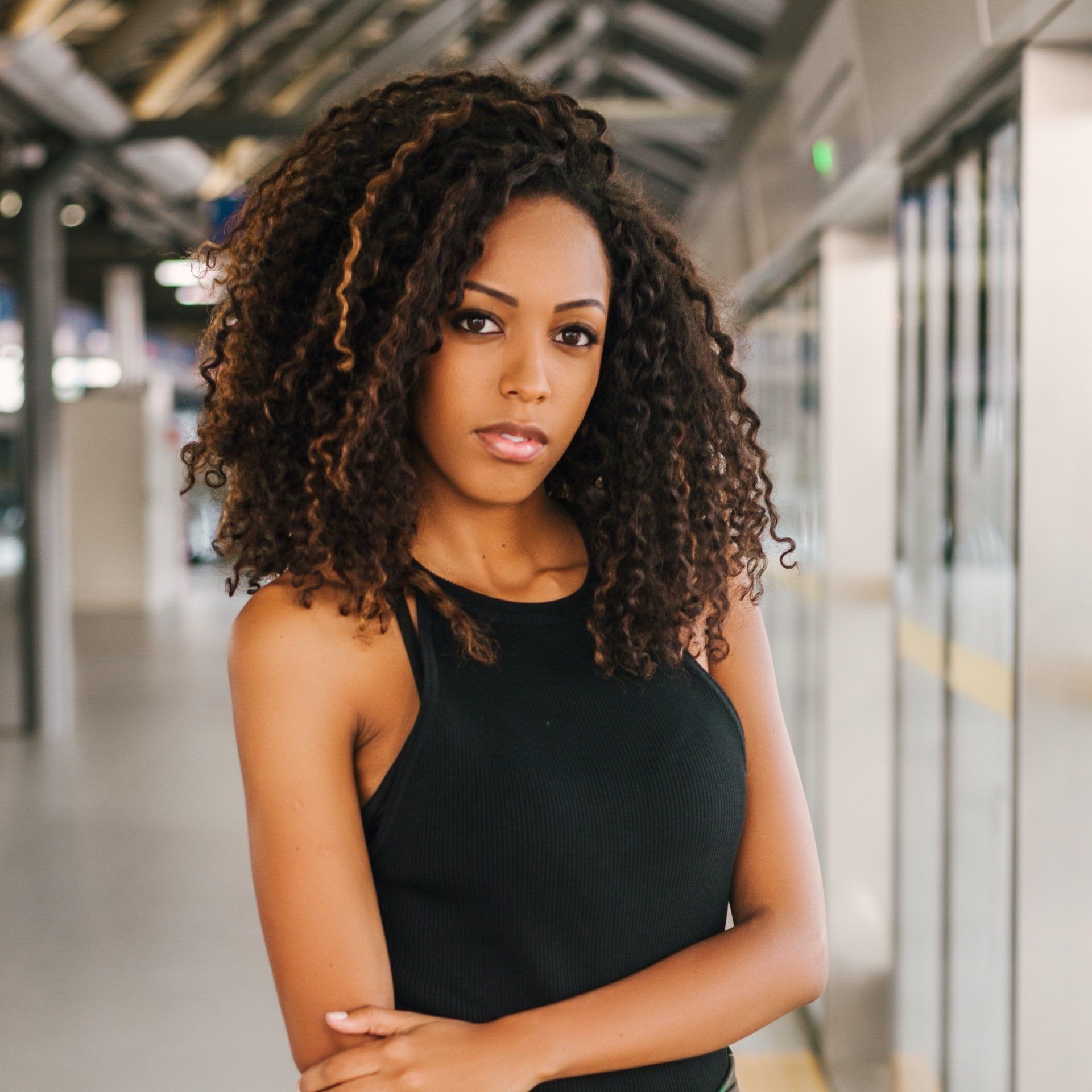 Woman with curly brown hair in black top, looking at the camera in a train station.