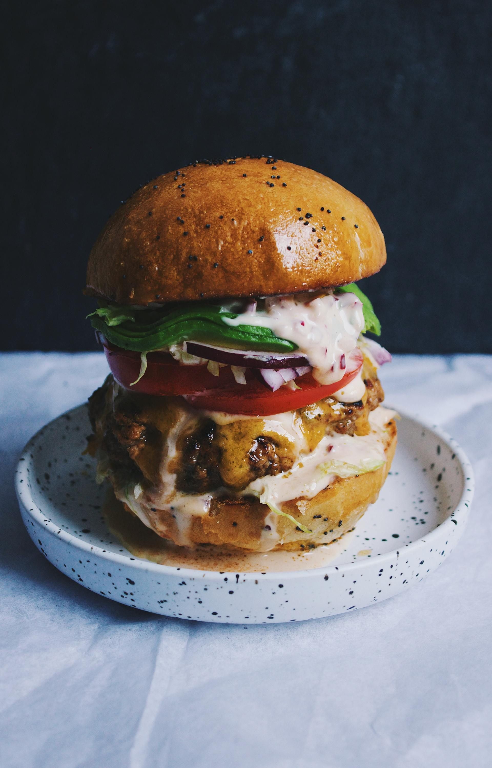 Tall stacked burger on a speckled plate against a dark background.