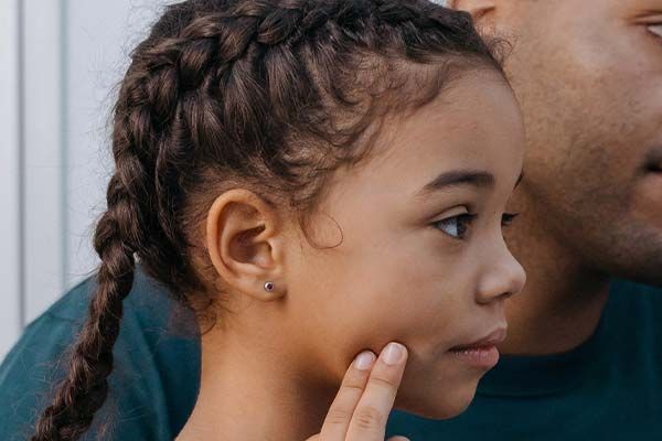 Young girl with braids, touched on the cheek, looks thoughtfully; man's arm and face partially visible.