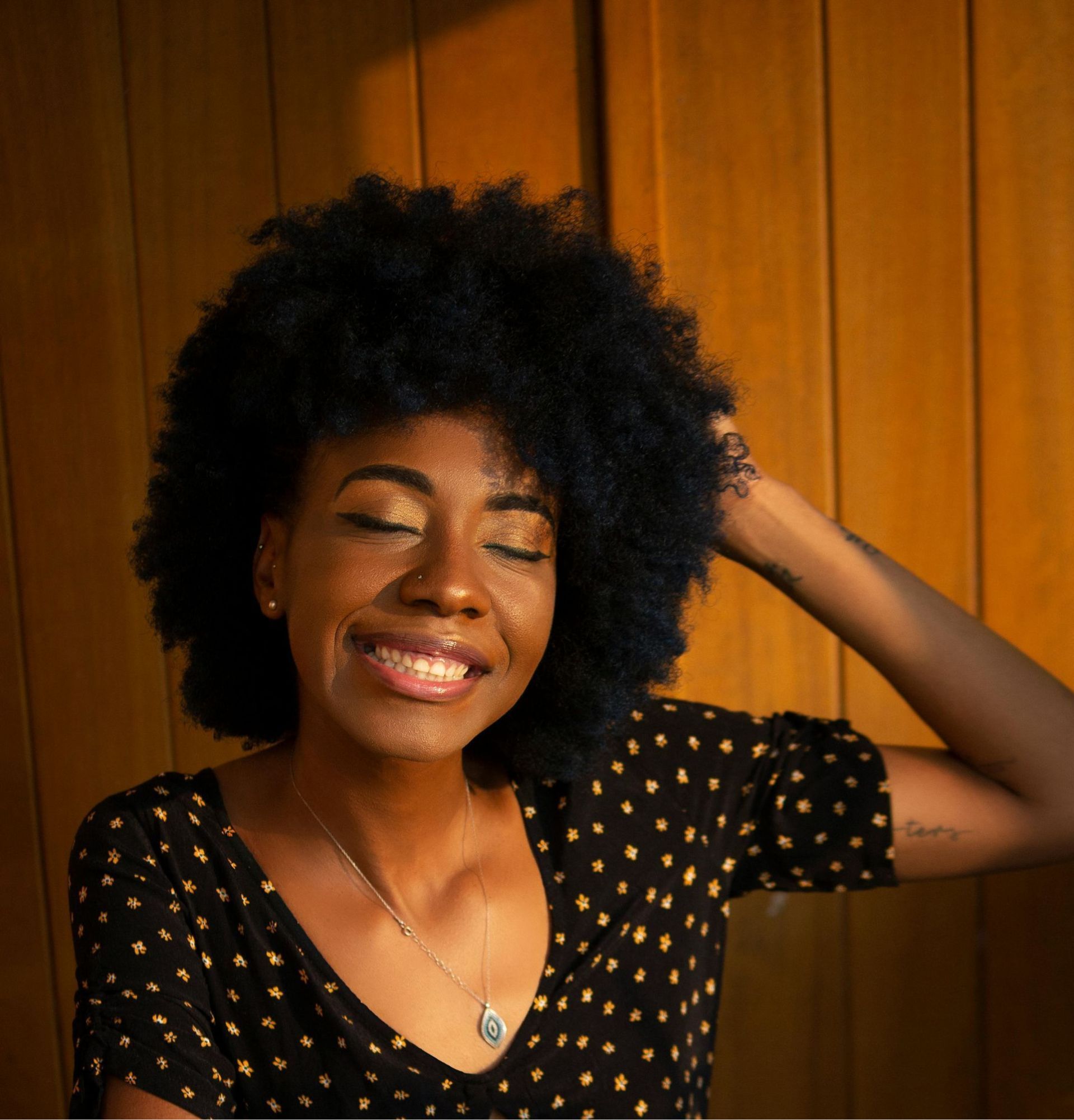 Woman with dark curly hair smiles, eyes closed, hand in hair, wearing polka dot top.
