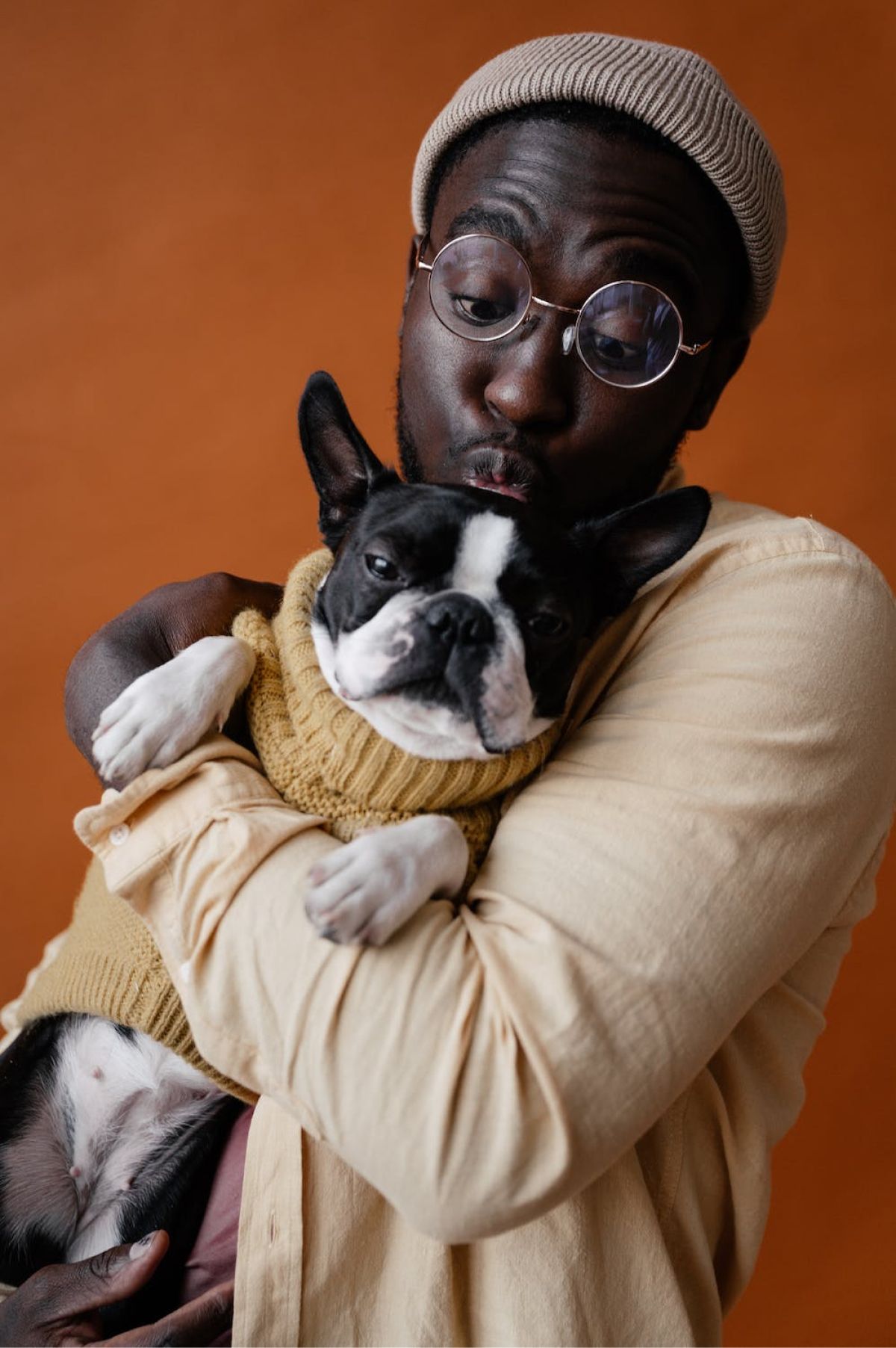 Mann med briller kysser en bostonterrier, begge iført brune gensere mot oransje bakgrunn.