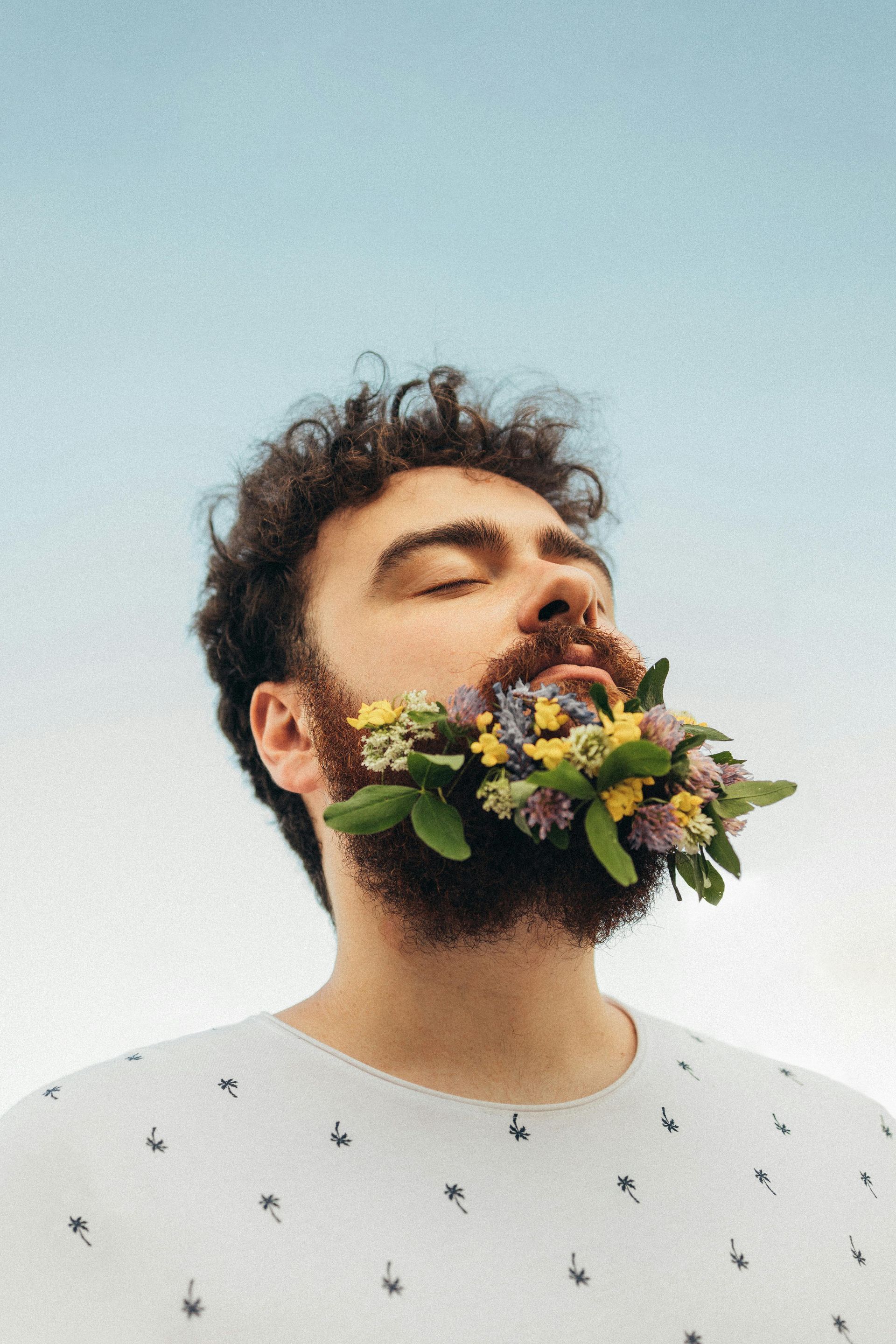 Mann med krøllete hår og skjegg fylt med fargerike blomster; øynene lukket, blå himmel i bakgrunnen.