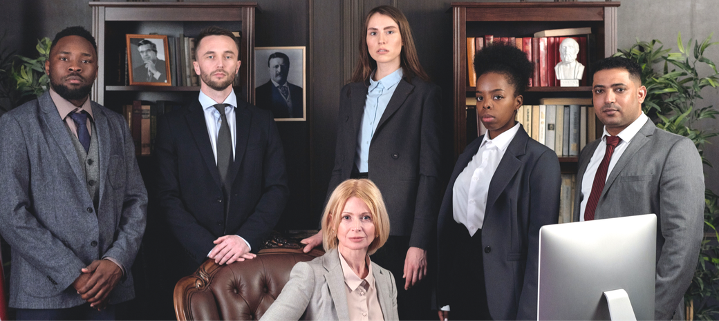 Group of seven people in suits, posing in an office setting with bookshelves and portraits in the background.
