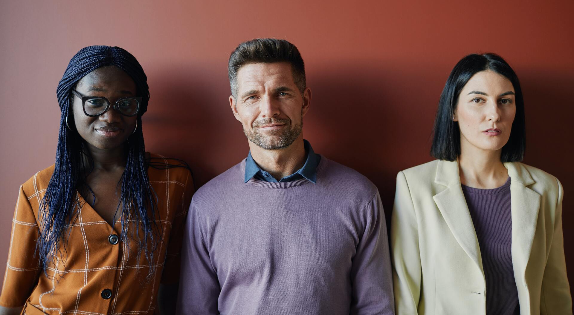 Three people stand against a maroon wall, looking toward the camera.