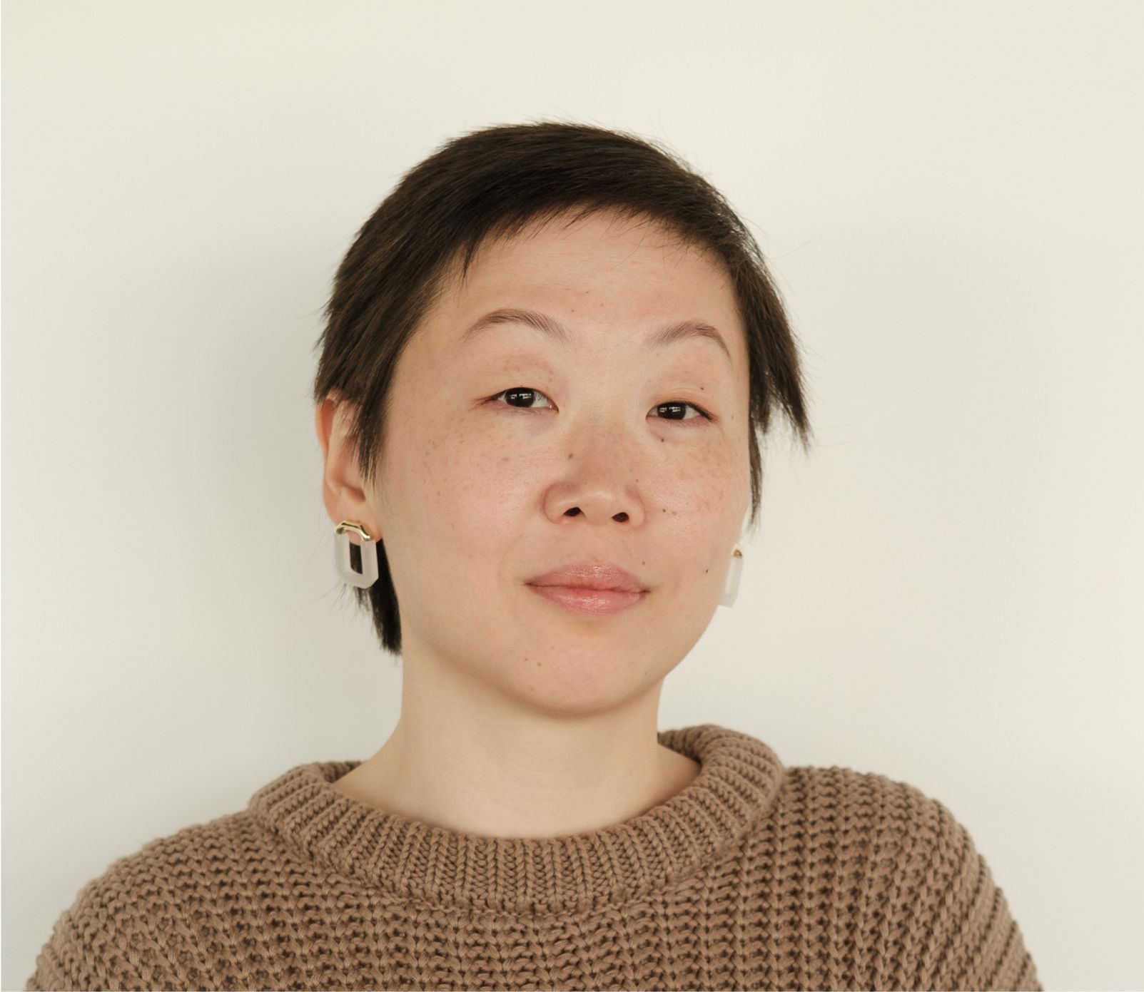 Woman with short dark hair, wearing a brown sweater and earrings, looking directly at the camera.