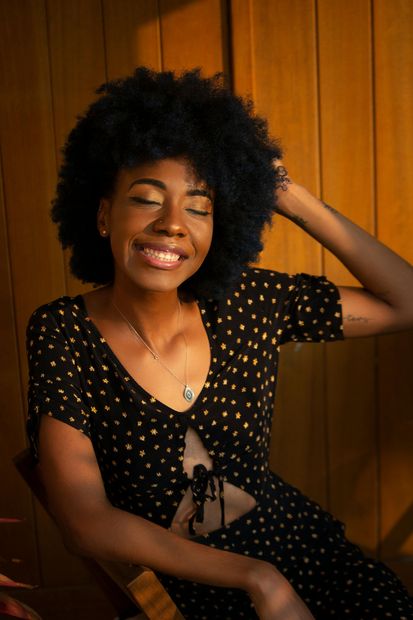 Smiling woman with curly hair in a black polka-dot dress, sitting against a wooden wall.