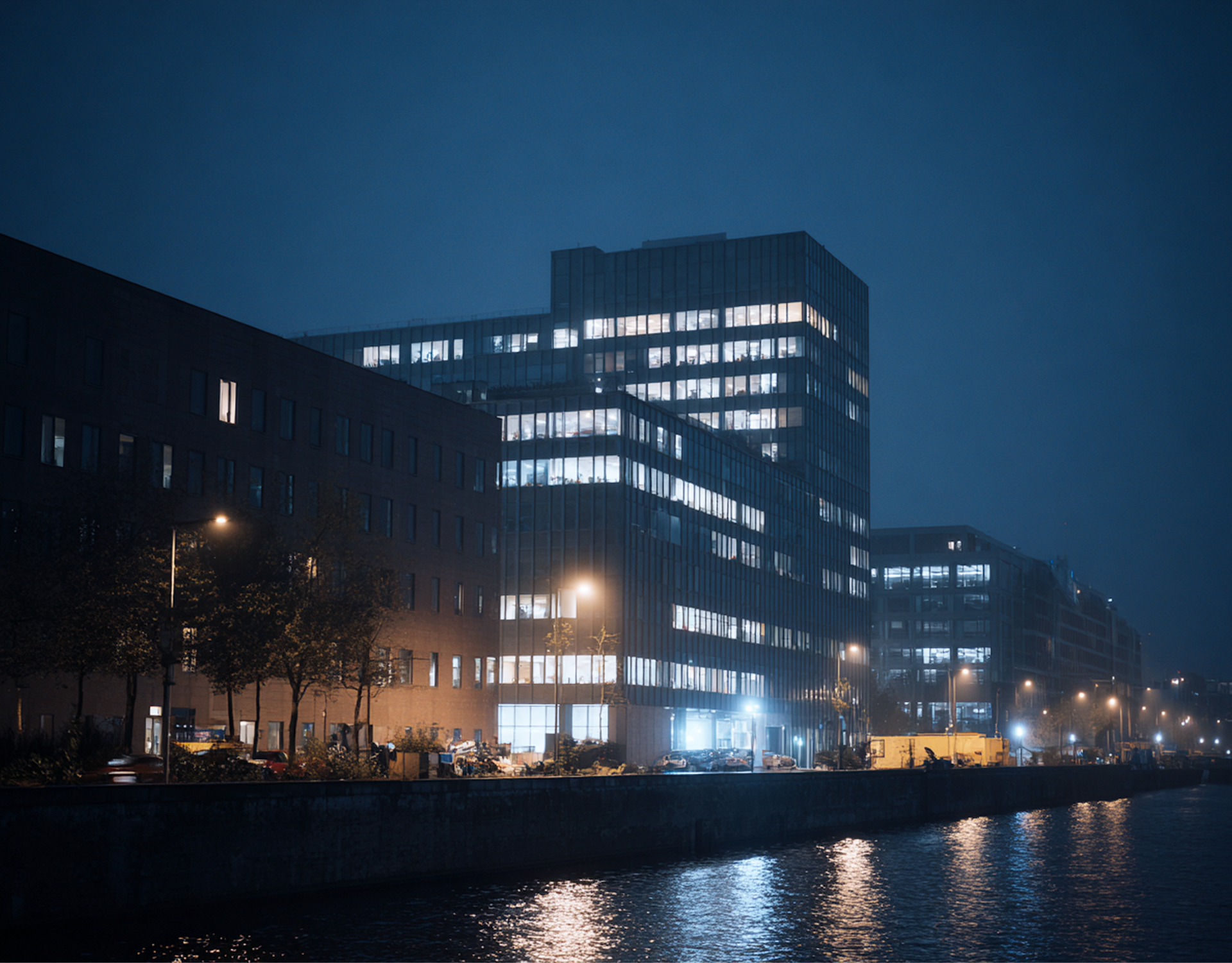 Grandi edifici per uffici illuminati di notte lungo la riva di un fiume, le cui luci brillanti si riflettono nell'acqua scura.